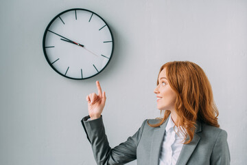  redhead businesswoman pointing with finger and looking at wall clock on grey