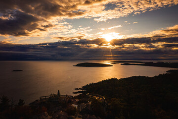 Beautiful sunrise aerial photograph above Sugarloaf Mountain looking towards Presque Isle peninsula and islands in Lake Superior with the sun shining between clouds and reflecting off of the water.