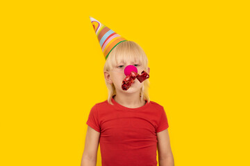 Child in festive hat with red clown nose and whistle. Festive mood. Portrait of boy on yellow...
