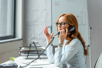  businesswoman gesturing and talking on landline telephone, while sitting at workplace on blurred background - obrazy, fototapety, plakaty