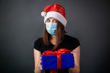 A woman in a Christmas cap and a medical mask holds a gift box in her hands while standing against a gray background.