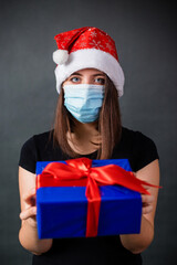 A woman in a Christmas cap and a medical mask holds a gift box in her hands while standing against a gray background.