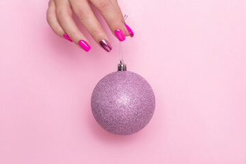 
Beautiful female hand with festive manicure nails holding Christmas toy, glitter ball on pink background