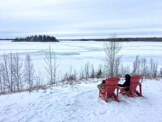 A mother and her daughter sitting in red adirondack chairs on a snowy winter day admiring the view at Elk Island Park, Alberta, Canada.