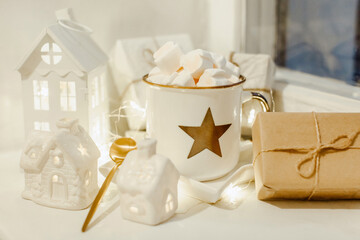 Christmas composition in light colors. White houses, gifts in white packages, festive lights, white cocoa mug with marshmelot and golden spoon. The concept of Christmas and New Year. Horizontal image.