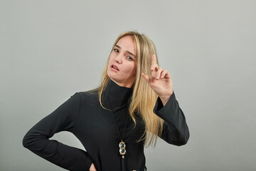 Hand points index finger up, pointing forefinger surprise gift option, great idea. Young attractive woman, dressed black sweater with green eyes, blonde hair, background