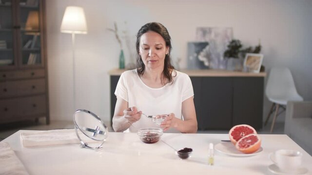Medium shot of middle-aged mixed race woman adding coconut sugar to bowl with mixed grapefruit pulp and coffee grounds mixing all ingredients together to make scrub