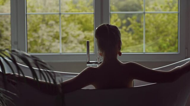 Back view of a woman relaxing in a bathtub with a large window
