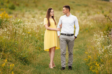 young couple in love look at each other in a grass field in sunny day. girl in a yellow dress and a guy with a stylish haircut. portrait of couple walking on countryside