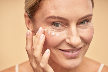Skin care. Close up of beautiful mature woman applying anti-aging cream under the eyes, isolated on beige background