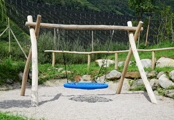 Vogelschaukel mit blauer Schaukel und Holzgerüst auf Sand auf einem Spielplatz mitten im Grünen - Spielgeräte im Freien