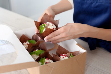 Closeup view of hands of female confectioner or baker packing tasty decorated muffins with cream top to delivery box. Ready cupcakes with berries and mint in package.