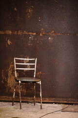 A rusty chair with peeling paint and a shabby leatherette seat against a rusty wall on the concrete floor.