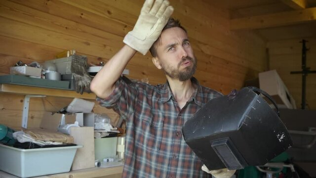 Portrait of tired craftsman removes welding mask after returning to workshop, different construction tools in the background. DIY concept.