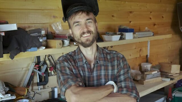 Portrait of bearded worker man removes welding mask after returning to workshop after work, different construction tools in the background. Cheerful craftsman. DIY concept.