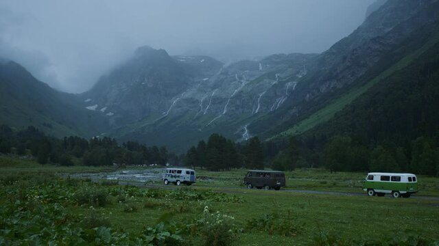 Four cars drive against the backdrop of mountains with a glacier and a waterfall. Powerful passable UAZ car rides on a bad road in a mountain valley. Slow motion