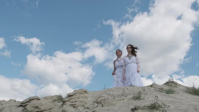 Two young girls in white Boho dresses pose together on a sand hill against a blue sky. Happy young people, teenage friendship