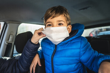 Portrait of small caucasian boy in car wearing protective face mask and his mother's hand helping him to adjust - covid-19 coronavirus new normal concept