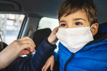Fototapeta na wymiar Portrait of small caucasian boy in car wearing protective face mask and his mother's hand helping him to adjust - covid-19 coronavirus new normal concept