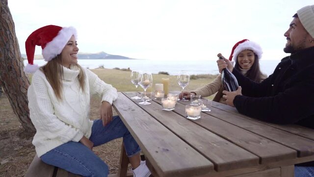 Three mixed race friends sitting on a table outdoors in Santa hats celebrating Christmas and new year holidays at sea resort in winter vacations - Funny scene of guy uncorking prosecco wine bottle
