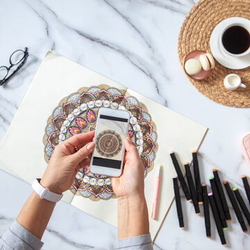 Woman is taking photos of colored mandala from sketchbook on white marble desk with displayed object