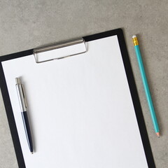 Template of white paper with a ballpoint pen and simple pencil on light grey concrete background in a black tablet with a clip. Stock photo with empty space for text