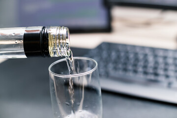 Drinking alcohol at work or office. Vodka is poured in a glass. Defocused keyboard and desk showing...