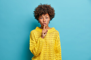 Surprised dark skinned young woman makes shush gesture asks to keep information in secret shows...