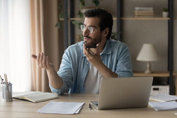 Obraz premium Young Caucasian man sit at desk in home office work on laptop record audio message on smartphone. Millennial male use computer speak talk on call activate digital voice assistant on cellphone.
