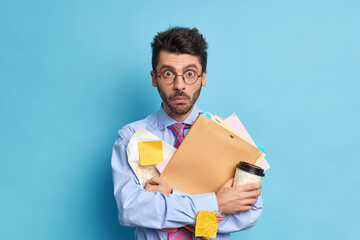 Shocked student with thick bristle prepares for studying courses holds papers and disposable cup of...
