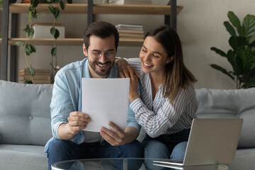 Happy young Caucasian couple feel overjoyed read good banking paper notice. Smiling millennial man...