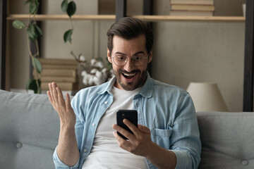 Stunned millennial male in glasses sit on sofa at home look at cellphone screen shocked by amazing news online. Amazed young Caucasian man surprised by unexpected good sale offer deal on smartphone.