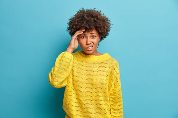 Displeased curly haired Afro American woman keeps hand on temple suffers migraine or severe headache frowns face and has problems wears casual knitted yellow jumper isolated over blue background