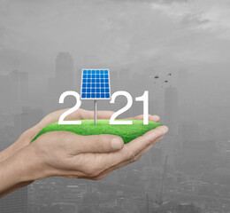 2021 white text with solar cell on green grass field in man hands over pollution city tower and skyscraper with birds, Happy new year 2021 ecological cover concept