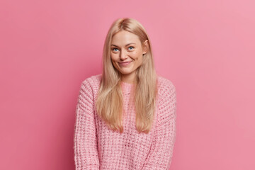 Fototapeta premium Cute pleased blonde woman smiles pleasantly has romantic face expression dimples on cheeks concentrated above wears warm knitted sweater isolated over pink background. Dreamy European girl indoor
