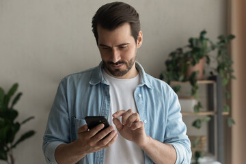 Young Caucasian man stand look at smartphone screen browse surf wireless internet on gadget. Serious millennial male use cellphone device, text or message online. Communication concept.