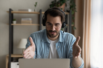 Young Caucasian man in headphones work online on laptop consult client on web. Serious millennial male in earphones have webcam digital talk or video call with business customer on computer.