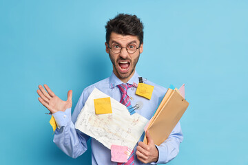 Angry annoyed man manager shouts with irritation being busy working and surrounded by papers and stickers of reminding dressed in formal clothes isolated on blue. Employee gives report of done work