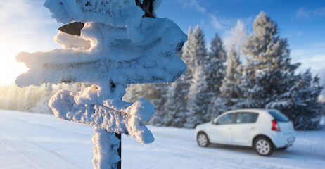 Fototapeta premium Snow covered signboard with wooden arrows on a background of a car on a road. Winter travel 