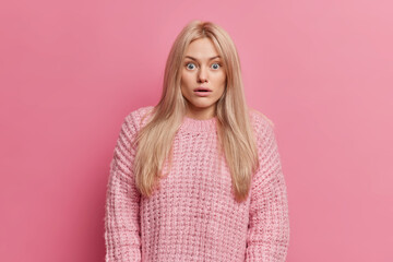 Shocked blonde young European woman has stupefied face expression stares bugged eyes cannot believe in shocking horrible news wears knitted sweater poses against pink background. Omg concept