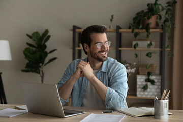 Smiling millennial Caucasian man sit at desk at home office distracted form computer job dreaming or thinking. Happy young male work online on laptop planning or pondering. Vision concept.