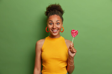 Happy millennial girl with curly hair wears yellow t shirt and earrings holds sweet lollipop poses against green background. Young dark skinned Afro American woman enjoys eating delicious candy