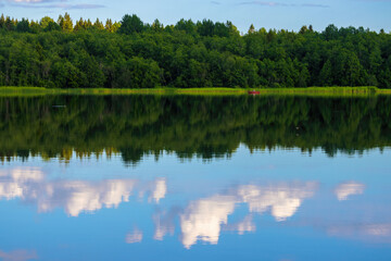 Obraz premium View of the lake and forest. Red boat in the distance. Outdoor recreation and fishing on the lake. Beautiful summer landscape. Clouds are reflected in the water. Travel and vacation in the countryside