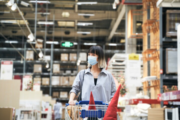 Asian woman wearing face mask push shopping cart in warehouse store buying Christmas decorations & gifts