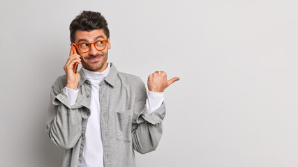 Pleased cheerful hipster guy has telephone conversation keeps modern smartphone near ear and points away on empty space wears glasses and shirt calls to friend isolated over grey background.