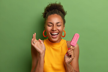 Overjoyed millennial woman with combed curly hair smiles broadly has fun and eats delicious...