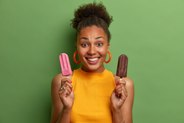 Photo of cheerful dark skinned African American woman holds strawberry and chocolate ice cream smiles broadly dressed in casual yellow t shirt poses against green background. People summer concept