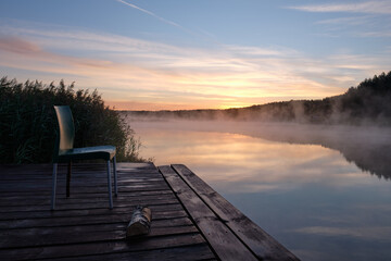 Summer, lake, early morning, beautiful sky, fog over the water. A wooden dock with an empty chair.