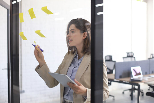 Attractive Busness Woman Post Stickers On Glass At Office And Writing On Them.