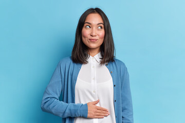 Pleased cheerful Asian woman keeps hand on belly feels full after delicious dinner dressed casually stands thoughtful against blue background. Happy young female finds out about her pregnancy © WHstudio Leushin N